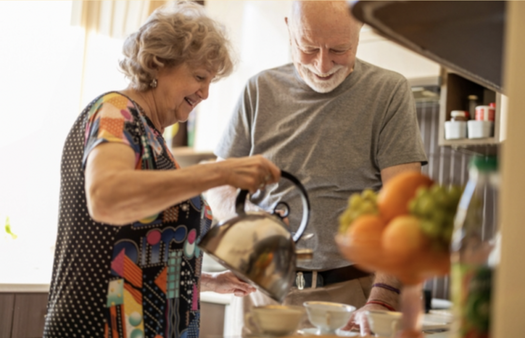 Mature couple smiling in their kitchen as the wife pours hot water into tea cups for herself and her husband