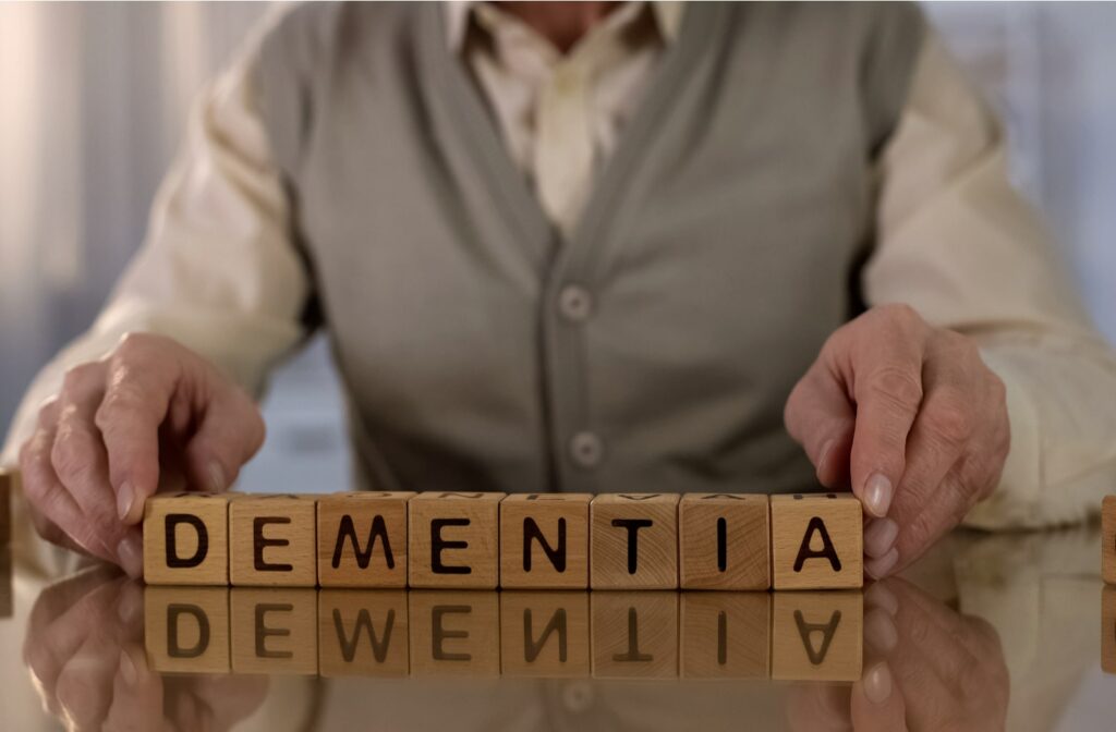Image of a mature man wearing a collared shirt and knitted vest placings his hands at both ends of the word dementia which is spelled out in block letters