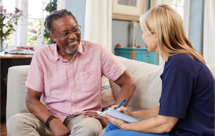 A female health aide sits with a smiling mature man during a care visit.