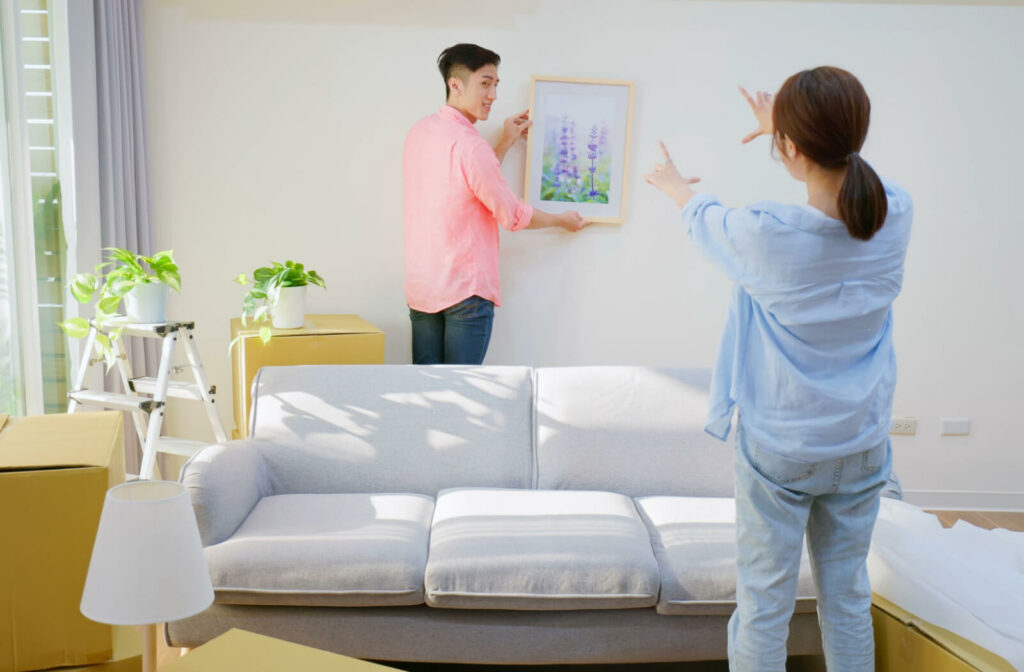 A male in a pink shirt and a female in a blue shirt is personalizing the room of their parent inside an assisted living facility. The man is holding a frame of a lavender flower on the wall while the woman is checking if the frame has the right angle. In the room, there is a grayish-white sofa and an indoor plant in the corner.