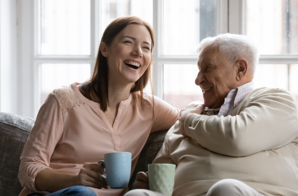A senior man and his daughter sitting on a couch smiling and talking to each other while holding a cup of tea.