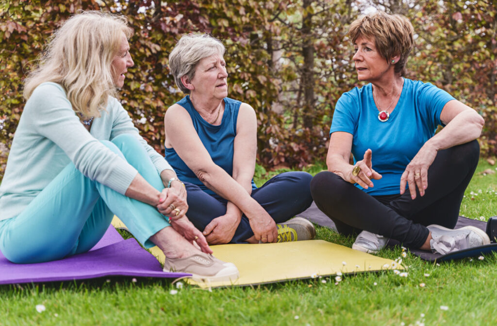 3 senior women sitting on yoga mats in a park and chatting.