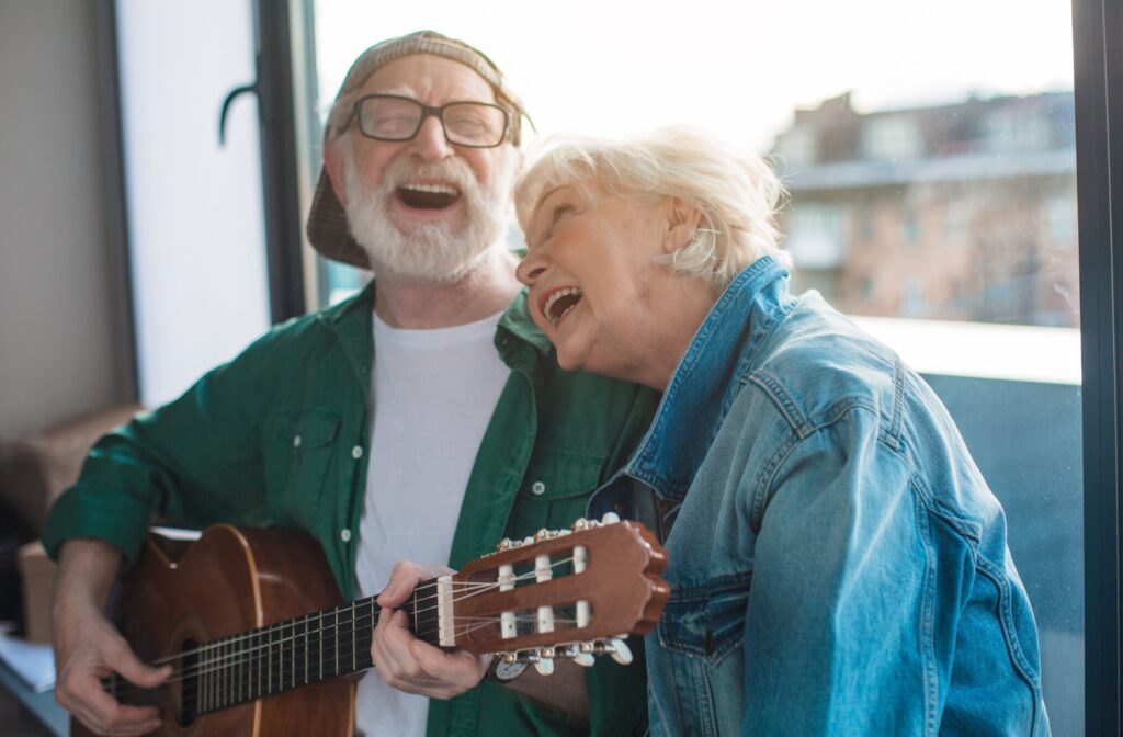 2 seniors sit on a couch in a well-lit room. Both are singing joyfully, 1 is playing guitar.