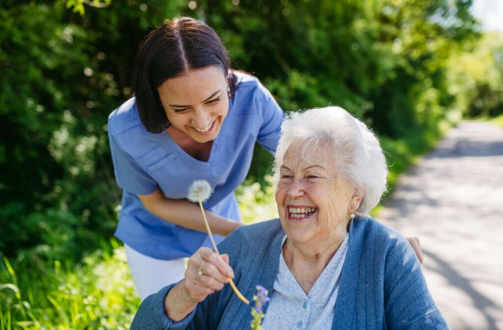 A senior woman with dementia smiling and holding up a dandelion while a caregiver pushes her wheelchair through a garden in memory care.