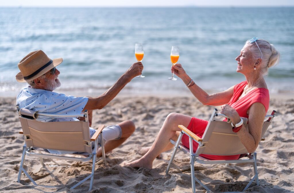 Two older adults enjoying a drink together while sitting in chairs on the beach.