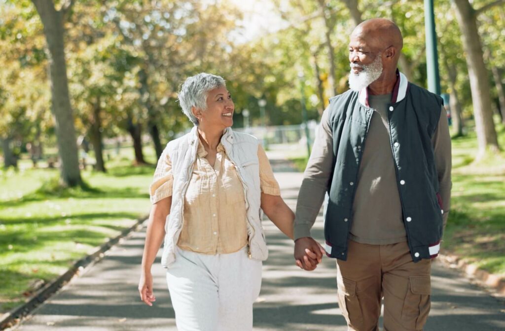 Two older adults holding hands while walking together on a tree-lined park path smiling and making eye contact