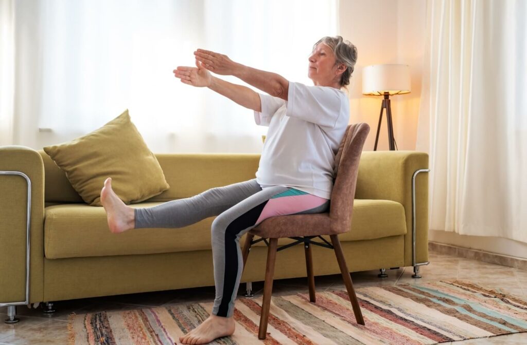 An older adult raises their arms and lifts their right leg while performing chair yoga in their living room