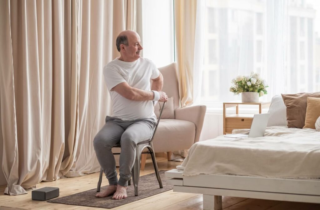 An older adult twists to one side while doing chair yoga in their sunlit bedroom
