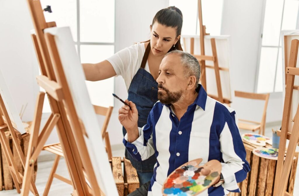 A senior in memory care takes an art class while getting help from their art teacher.