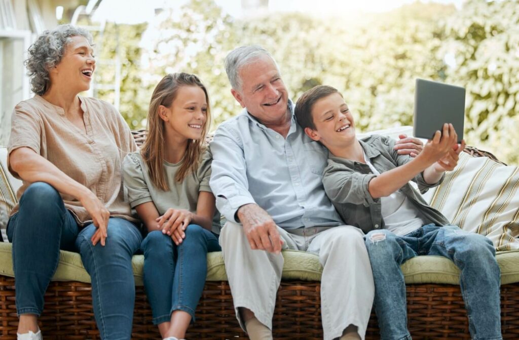 A grandchild holds up a tablet and laughs while their sibling and grandparents look at the screen during a visit in assisted living