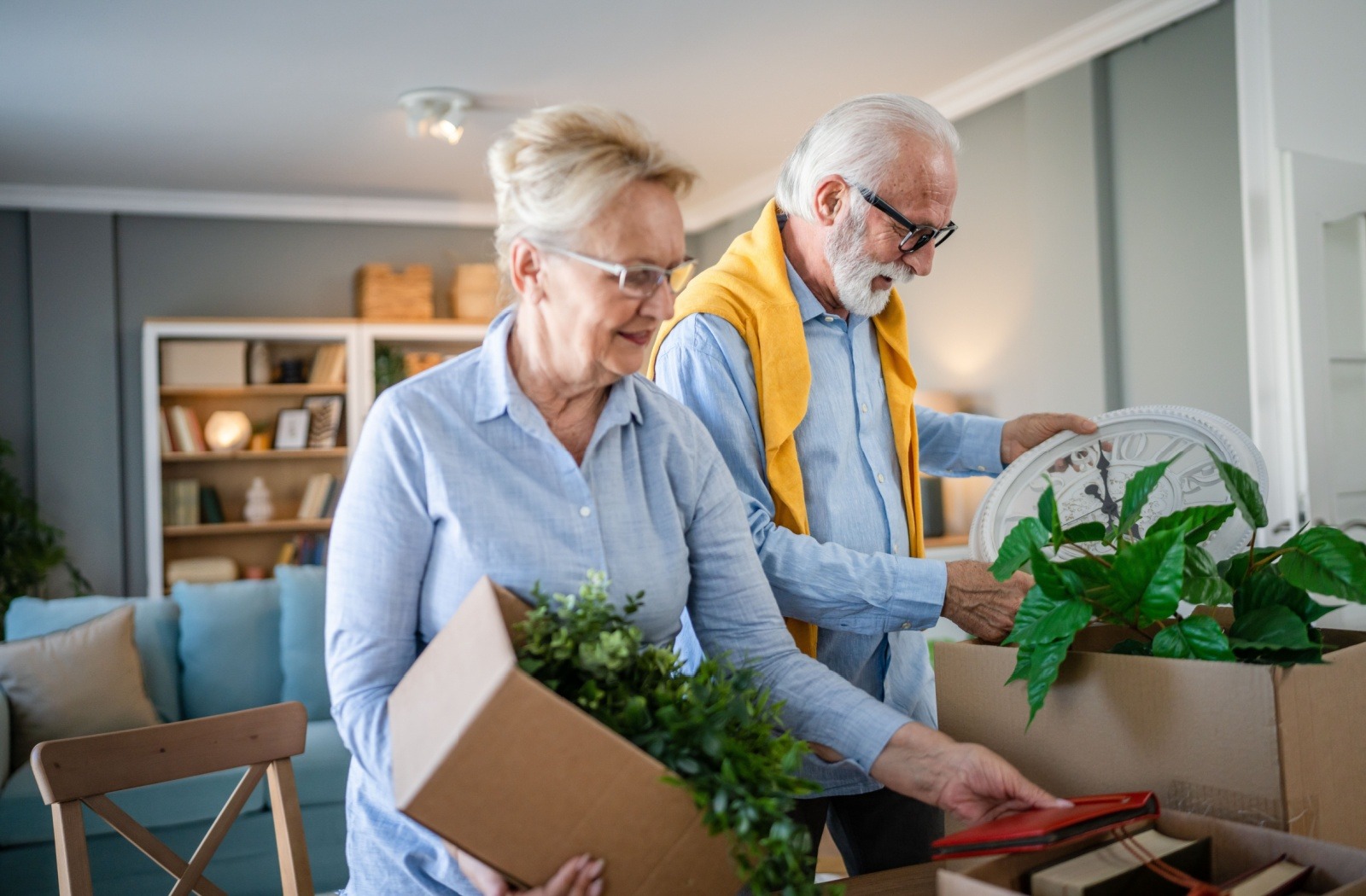 A senior couple packs some artificial houseplants into cardboard boxes as they prepare to downsize their home.