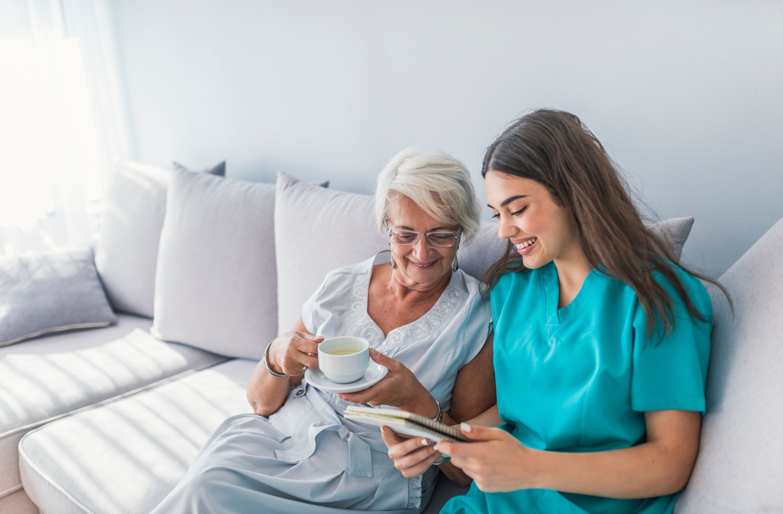 A senior drinks a cup of tea as a smiling caregiver reads some notes on a notepad.
