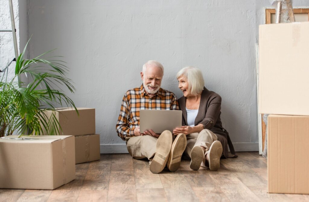 An excited senior couple sit on the floor surrounded by moving boxes, as they browse information on a laptop