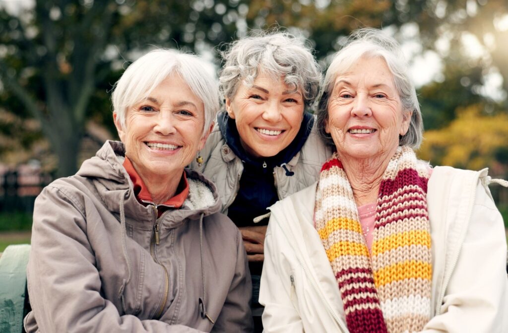 A group of 3 senior friends smile as they sit on an outdoor park bench on a brisk day
